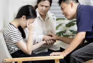 Asian mother and father looking kindly at daughter who has her face in her hands.