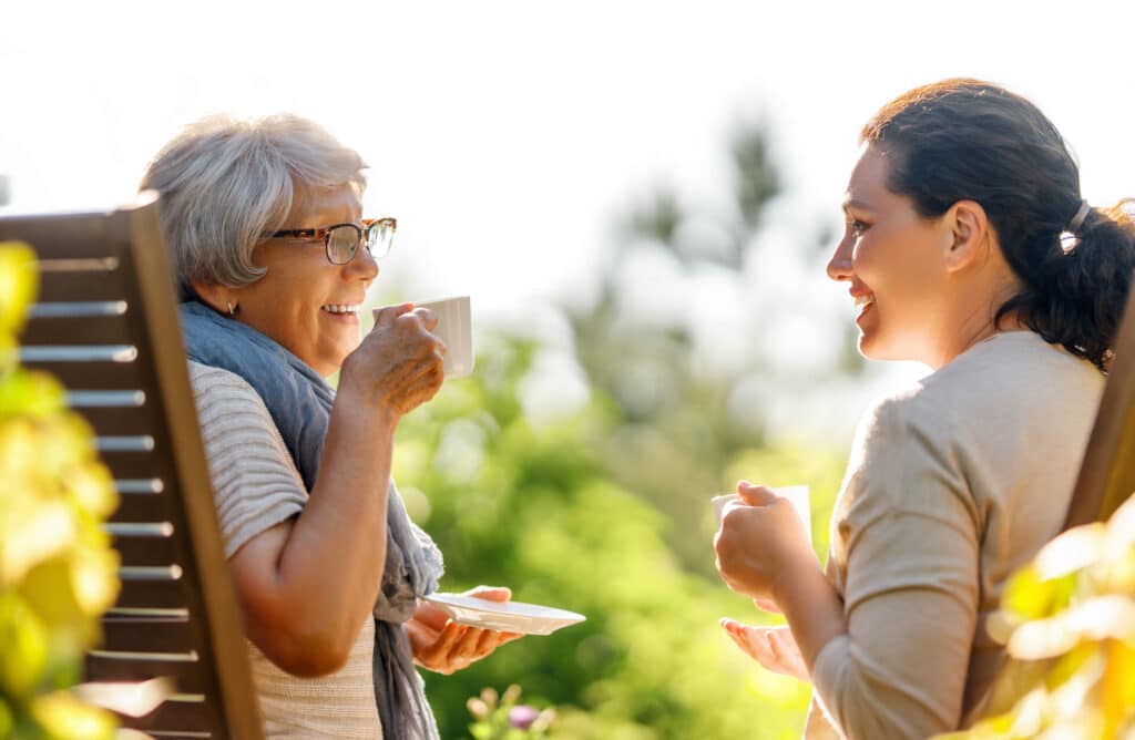 Happy young woman and her mother drinking tea in summer morning. Family sitting in the garden with cups and enjoying the conversation.