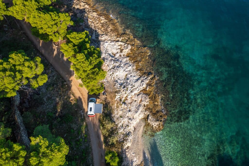 Aerial view of solitary camper parked on coastal road between Mediterranean forest and crystal-clear Adriatic waters along rocky shoreline.
